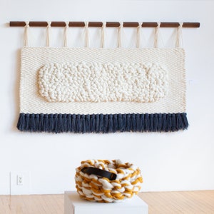 May include: A white wall tapestry with a textured oval design and a blue fringe. A yellow and white knitted basket with a black leather handle sits on a white pedestal.