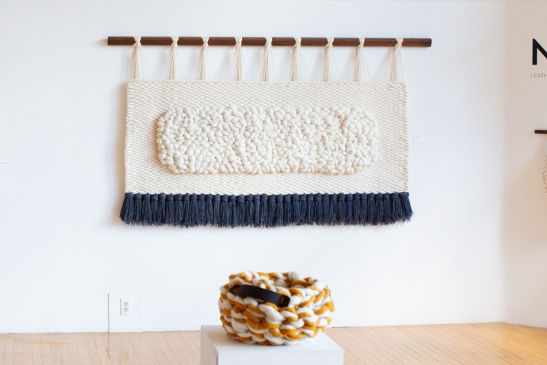 May include: A white wall tapestry with a textured oval design and a blue fringe. A yellow and white knitted basket with a black leather handle sits on a white pedestal.
