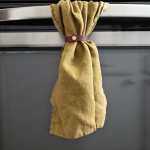 Olive green kitchen towel held by a rich wine-colored leather towel holder with brass hardware, displayed on a stainless steel oven handle. The pairing gives a warm, modern accent with subtle retro undertones.