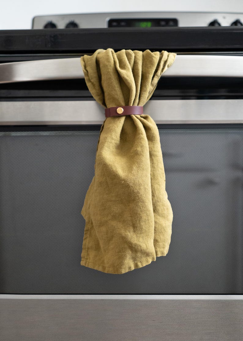 Olive green kitchen towel held by a rich wine-colored leather towel holder with brass hardware, displayed on a stainless steel oven handle. The pairing gives a warm, modern accent with subtle retro undertones.