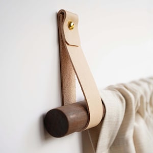 Könnte beinhalten: Ein hellfarbiger Lederriemen mit einem goldfarbenen Niet ist an einer dunkelbraunen Holzstange befestigt. Die Stange hält einen beigen Stoff. Der Lederriemen ist an einer weißen Wand befestigt.
