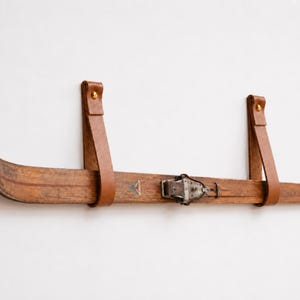 Op de afbeelding: Een vintage houten ski met leren riemen voor wandmontage. De ski heeft een metalen punt en een metalen binding. De leren riemen zijn bruin en bevestigd met messing klinknagels. Een uniek stuk rustieke decoratie.