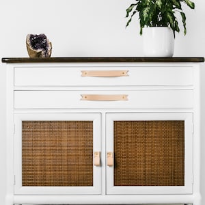 Keyaiira Veg Tan genuine leather cabinet and drawer handles with V-shaped pointed ends and polished brass mounting hardware installed on a white storage cabinet with woven cane doors, styled with greenery and decorative accents for a warm modern interior