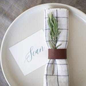 May include: A white linen napkin with a black and white checkered pattern is tied with a brown leather napkin ring. A sprig of greenery is tucked into the napkin. A white card with the name "Sean" written in blue ink is placed next to the napkin.