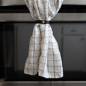White kitchen towel with black grid pattern neatly gathered by a black leather towel holder with brass hardware on a stainless steel oven handle.