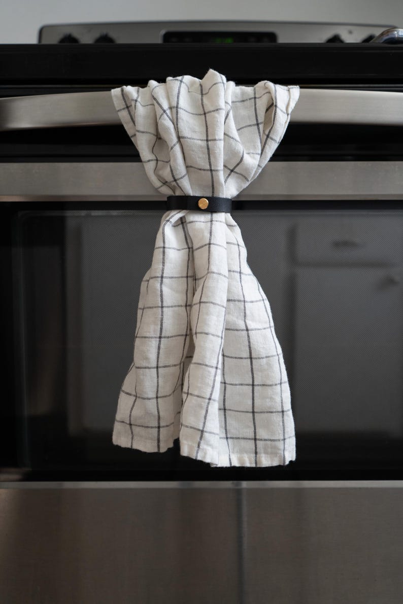 White kitchen towel with black grid pattern neatly gathered by a black leather towel holder with brass hardware on a stainless steel oven handle.