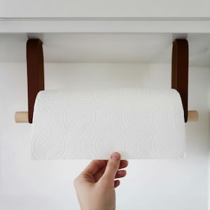 以下が含まれることがあります： 茶色の革と木製のペーパータオルホルダー。ホルダーは白いキャビネットに取り付けられています。繰り返しのパターンを持つ白いペーパータオルのロールがホルダーにあります。ホルダーは、茶色の革ストラップでキャビネットから吊り下げられています。