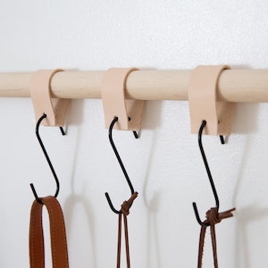 May include: Three leather and metal S-hooks hanging from a wooden rod. The hooks are black, and the leather is a natural tan color. The hooks are holding brown leather straps.