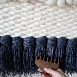 May include: A close-up of a hand holding a wooden comb with five tines. The comb is being used to brush the fringe of a woven wall hanging. The wall hanging is made of white and blue wool yarn.
