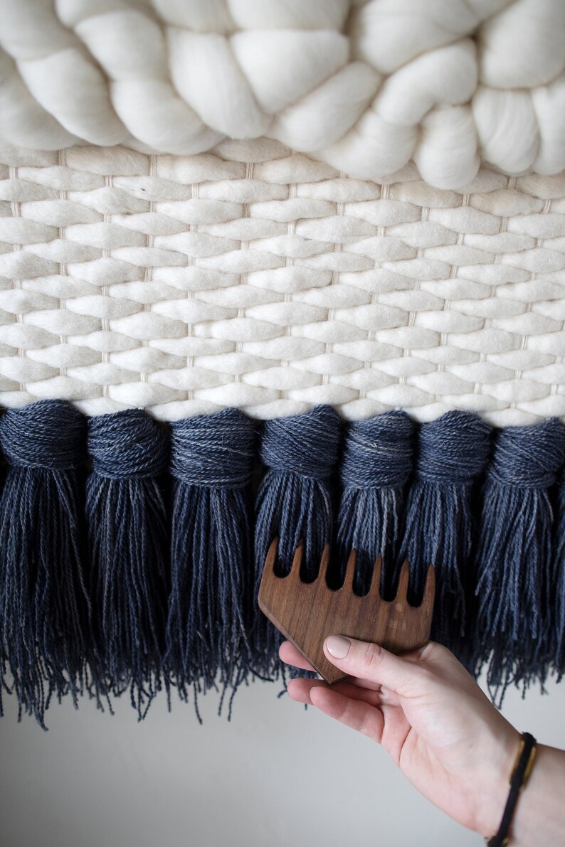 May include: A close-up of a hand holding a wooden comb with five tines. The comb is being used to brush the fringe of a woven wall hanging. The wall hanging is made of white and blue wool yarn.