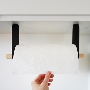 Könnte beinhalten: Ein schwarzer Papierhandtuchhalter mit einem natürlichen Holzdübel und schwarzen Lederriemen. Der Halter ist unter einem weißen Schrank montiert. Eine Rolle weißer Papierhandtücher befindet sich im Halter.