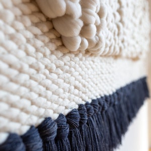May include: A white woven wall hanging with a textured, puffy design and a dark blue fringe border.