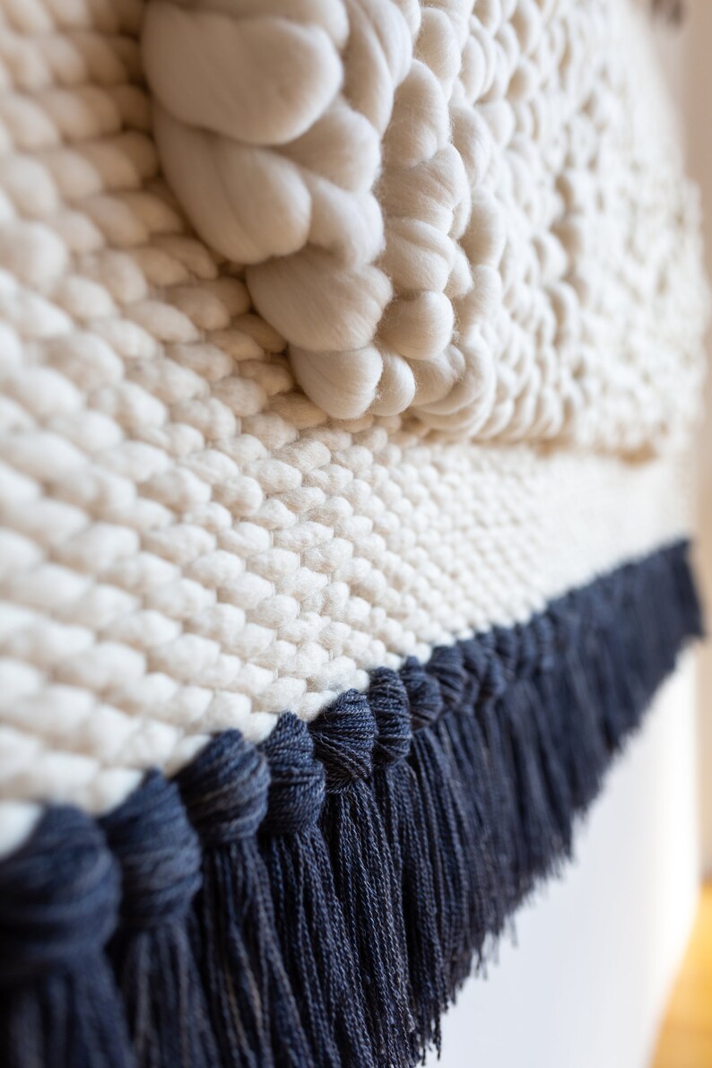 May include: A white woven wall hanging with a textured, puffy design and a dark blue fringe border.