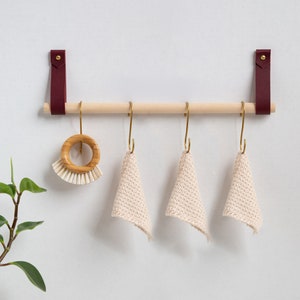 May include: A wooden bar with burgundy leather straps hangs on a white wall. Three brass hooks with cream-colored crocheted dishcloths hang from the bar. A wooden brush with white bristles is attached to one of the hooks.