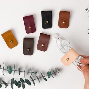 May include: A collection of leather earbud holders in various colors, including tan, brown, black, and burgundy, each with a gold-colored snap closure. A light tan leather holder with earbuds is held by a hand.