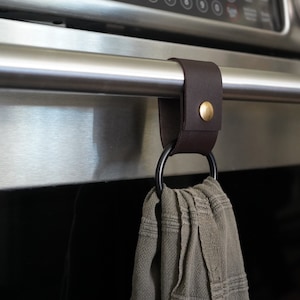 May include: A dark brown leather towel holder with a brass button is attached to a stainless steel oven handle. A gray towel hangs from a black metal ring. The oven display shows the time.