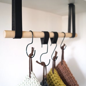 May include: A wooden kitchen rack with black metal hooks and leather straps. The rack is suspended from a white shelf and holds several crocheted pot holders in various colours. The wooden rod is light-coloured.