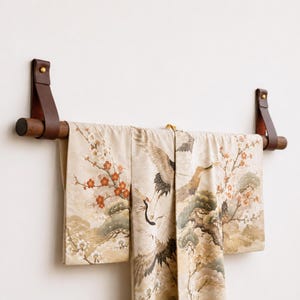 May include: A dark wood wall-mounted display rod suspended by two brown leather straps with brass hardware. A cream-coloured textile featuring a printed pattern of cranes, pine trees, and orange blossoms hangs draped over the horizontal wooden rod.