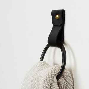 May include: A black leather towel ring with a brass rivet is mounted on a white wall. A black metal ring holds a folded, light-colored, textured towel. The leather strap is curved and attached to the wall.