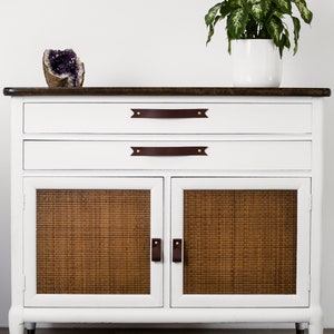 Keyaiira Mahogany genuine leather cabinet and drawer handles with V-shaped pointed ends and polished brass mounting hardware installed on a white storage cabinet with woven cane doors, styled with greenery and decor to showcase the rich warm leather color