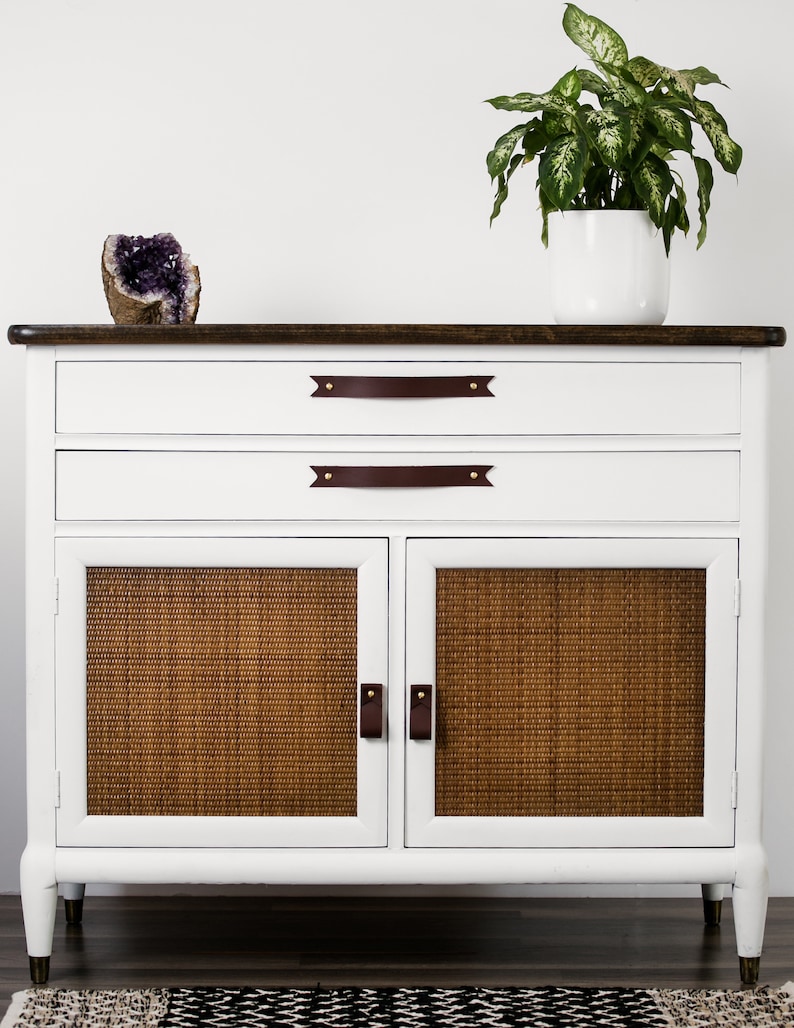 Keyaiira Mahogany genuine leather cabinet and drawer handles with V-shaped pointed ends and polished brass mounting hardware installed on a white storage cabinet with woven cane doors, styled with greenery and decor to showcase the rich warm leather color