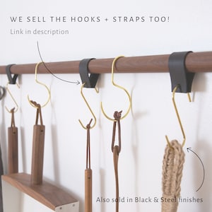 May include: A wooden wall mounted rod with black leather straps and gold metal hooks. The hooks are hanging from the rod and have brown leather straps attached to them. The text on the image reads: 'WE SELL THE HOOKS + STRAPS TOO! Link in description Also sold in Black & Steel finishes'.