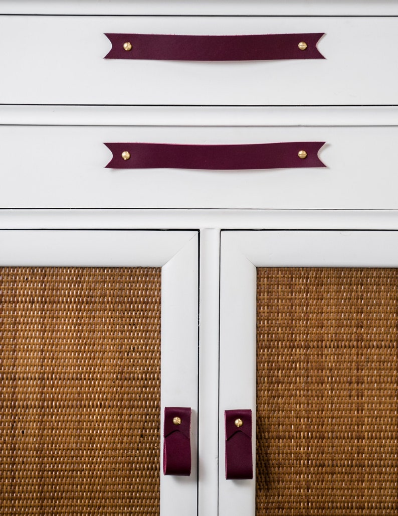 Keyaiira Wine genuine leather drawer handle with V-shaped pointed ends and polished brass mounting hardware installed horizontally on a white cabinet above woven cane doors, highlighting the deep Wine leather color, natural texture and handcrafted furniture hardware