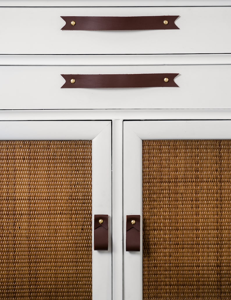 Keyaiira Chocolate genuine leather drawer handle with V-shaped pointed ends and polished brass mounting hardware installed horizontally on a white cabinet above woven cane doors, highlighting the rich Chocolate leather tone and handcrafted raised grip