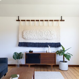 May include: A white and blue woven wall hanging with a fringe border hangs above a mid-century modern style wooden cabinet with black doors. A houseplant sits on the floor next to the cabinet.
