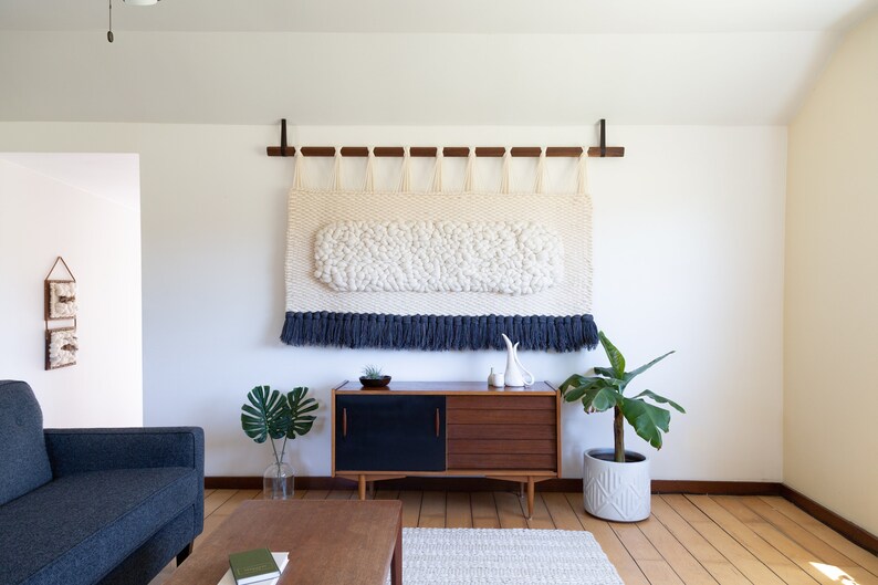 May include: A white and blue woven wall hanging with a fringe border hangs above a mid-century modern style wooden cabinet with black doors. A houseplant sits on the floor next to the cabinet.