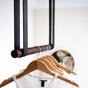 May include: A contemporary clothes rail featuring a dark brown wooden bar and black straps hanging from the ceiling. Several wooden hangers hold white tops. The design is minimalist and functional, suitable for a bedroom or closet.