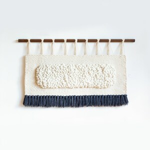 May include: A white woven wall hanging with a raised oval design and a black fringe border. The hanging is attached to a wooden dowel rod.