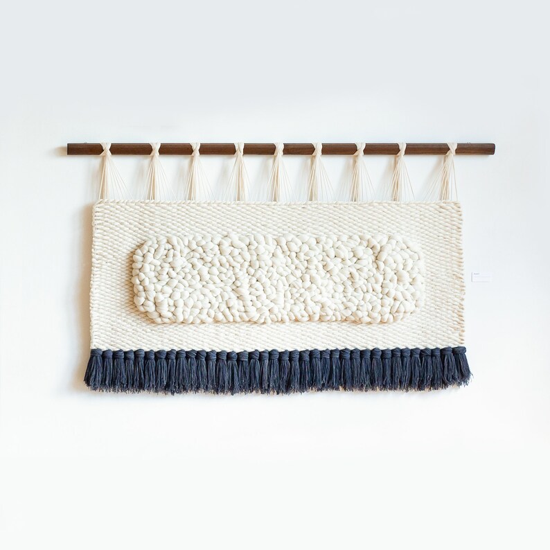 May include: A white woven wall hanging with a raised oval design and a black fringe border. The hanging is attached to a wooden dowel rod.