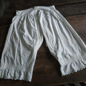 May include: A pair of white cotton bloomers featuring a gathered waistband and wide legs that taper towards the bottom. The hem of each leg is finished with a band of intricate smocking detail and a scalloped lace edge trim along the bottom.