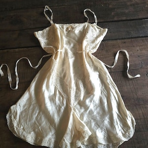 May include: A cream-colored satin slip dress featuring delicate lace trim along the neckline, bodice, and hem. The garment includes thin spaghetti straps and a matching satin tie belt at the waist, creating a soft, flowing silhouette with intricate lace detailing.