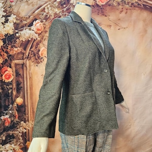May include: A charcoal grey blazer with a textured weave, featuring a notched lapel, two front buttons, and two patch pockets. The blazer is worn over a light grey sweater and paired with plaid trousers, showcasing a structured, long-sleeved design.