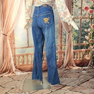May include: High-waisted blue denim jeans featuring a wide-leg silhouette and a yellow floral embroidery detail on the back pocket. The jeans have a classic five-pocket design with a dark wash finish and a visible brand label on the waistband.