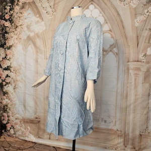 May include: A light blue vintage coat with a floral pattern. The coat features a high collar, three-quarter sleeves, and button closures down the front. The fabric has a subtle sheen, and the coat is displayed on a mannequin.