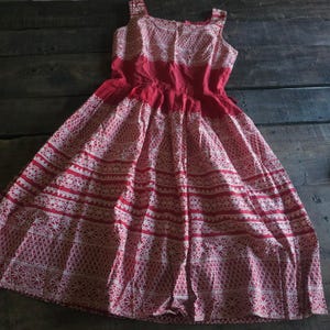 May include: A sleeveless red dress featuring a square neckline and a full, pleated skirt. The fabric is decorated with a repeating white geometric lace-like pattern arranged in horizontal bands across the bodice and the skirt.