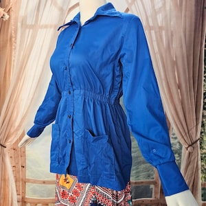 May include: A vibrant blue long-sleeve button-down shirt featuring a classic collar, a cinched elastic waistline, and two front patch pockets. The garment has long sleeves with fitted cuffs and a button-up front closure, displayed on a mannequin.