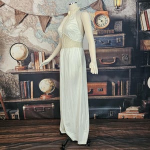 May include: A vintage ivory halter dress on a mannequin. The dress features a crochet bodice and a flowing skirt. The neckline and waist have crochet detailing. The dress is floor-length and appears to be made of a silky material.