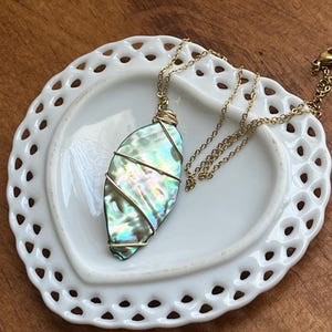 May include: Abalone shell pendant necklace. The iridescent, oval-shaped shell is wrapped in gold-colored wire and suspended from a delicate gold chain. The necklace rests on a white, heart-shaped dish with a decorative, openwork border. The background is a wooden surface.