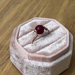 May include: A gold-colored wire ring featuring a deep red, round gemstone. The ring is displayed inside a pink velvet jewelry box with a hexagonal shape. The box has a soft, plush texture and a ribbed border.
