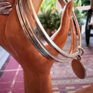May include: Silver bangles with a small, round charm hanging from one. The bangles are displayed on a wooden hand-shaped jewelry holder. The jewelry holder is a light brown color. The background includes a patterned rug and a glimpse of a chair.