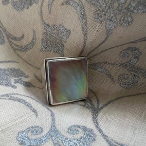 May include: A square silver ring featuring a large, iridescent, rectangular stone. The stone displays a mix of colours, including green, purple, and gold. The ring is set against a patterned, neutral-toned fabric.