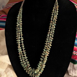 May include: A turquoise-colored necklace with a multi-strand design. The necklace is composed of small, irregularly shaped turquoise beads strung together with a silver-colored material. The necklace is displayed on a black mannequin.