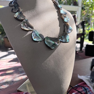 May include: A statement necklace featuring iridescent, shell-shaped pendants. The pendants display a range of colors, including blues, greens, and purples, and are strung together to form a unique piece of jewelry. The necklace is displayed on a mannequin.