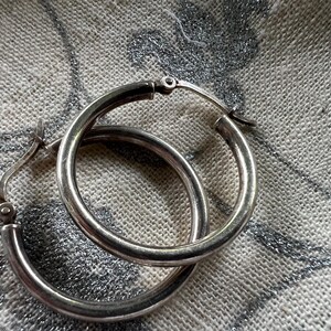 May include: A pair of silver hoop earrings. The earrings are round and have a simple, classic design. They are made of a shiny, metallic material and are shown against a textured, light-colored background.