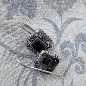 May include: A pair of silver-toned earrings with a rectangular shape and a central black stone. The earrings have a decorative, ornate design around the stone and a hook closure. The earrings are displayed on a patterned, light-colored fabric.
