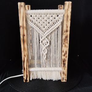 Op de afbeelding: Een witte macramé lampenkap met een geometrisch patroon hangt aan een houten frame. Het frame heeft een rustieke, gebrande houten afwerking.